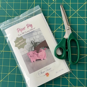 May include: A clear plastic bag containing a Piper Pig oven mitt sewing pattern with a photo of a pink pig-shaped mitt, alongside a pair of green-handled metal scissors. The pattern includes text and a video tutorial link, labeled SKU: PO-104.