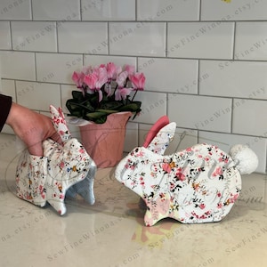 Peut inclure: Deux maniques en forme de lapin avec des motifs floraux. L'une est tenue par une main. Les maniques ont un tissu blanc avec des fleurs roses et rouges. Les oreilles sont roses et l'une a une queue en pompon blanc. Un pot de fleurs rose avec des fleurs roses est en arri&egrave;re-plan.