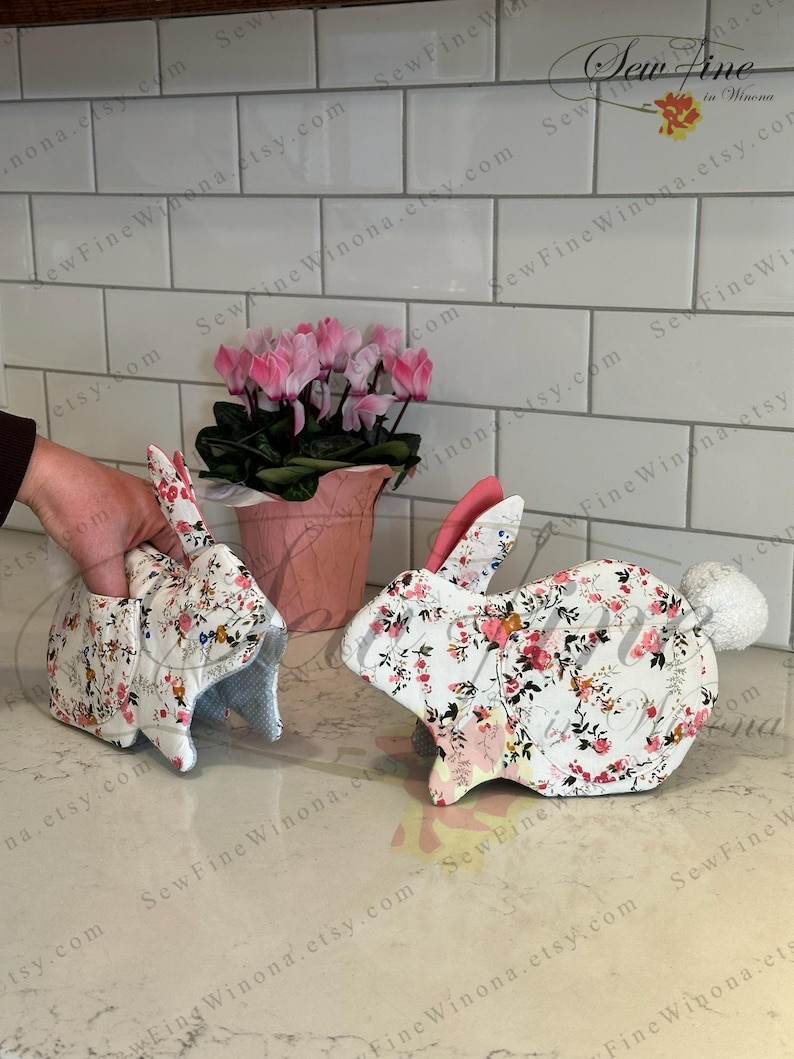 Peut inclure: Deux maniques en forme de lapin avec des motifs floraux. L'une est tenue par une main. Les maniques ont un tissu blanc avec des fleurs roses et rouges. Les oreilles sont roses et l'une a une queue en pompon blanc. Un pot de fleurs rose avec des fleurs roses est en arri&egrave;re-plan.
