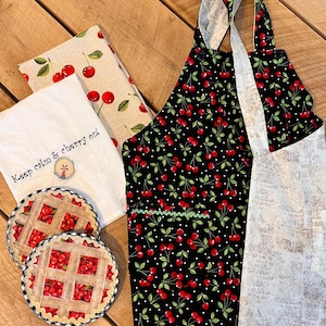 May include: A kitchen set featuring a black apron with a red cherry pattern, matching tea towels, and cherry pie-themed coasters. One tea towel has the text "Keep calm & cherry on!" The set is arranged on a wooden surface.