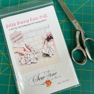 May include: A sewing pattern for a Betsy Bunny Oven Mitt in a clear plastic bag, featuring a photo of floral bunny-shaped oven mitts, the text 'Video tutorial link included!', and 'Sew Fine in Winona' branding with SKU: BO-102, next to silver scissors.