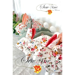 Peut inclure: Une collection de peluches lapins faites &agrave; la main avec du tissu floral et des couleurs d'oreilles contrast&eacute;es. Les lapins ont des queues en pompons blancs et sont dispos&eacute;s en rang&eacute;e. Le texte "Sew Fine in Winona" est visible.