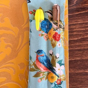 Puede incluir: Un estuche de tela azul claro con un estampado floral y de p&aacute;jaros, que contiene un rotulador amarillo y un bol&iacute;grafo de madera. El estuche est&aacute; decorado con flores y p&aacute;jaros de colores. El texto "Birds and Blooms" est&aacute; en la parte superior.