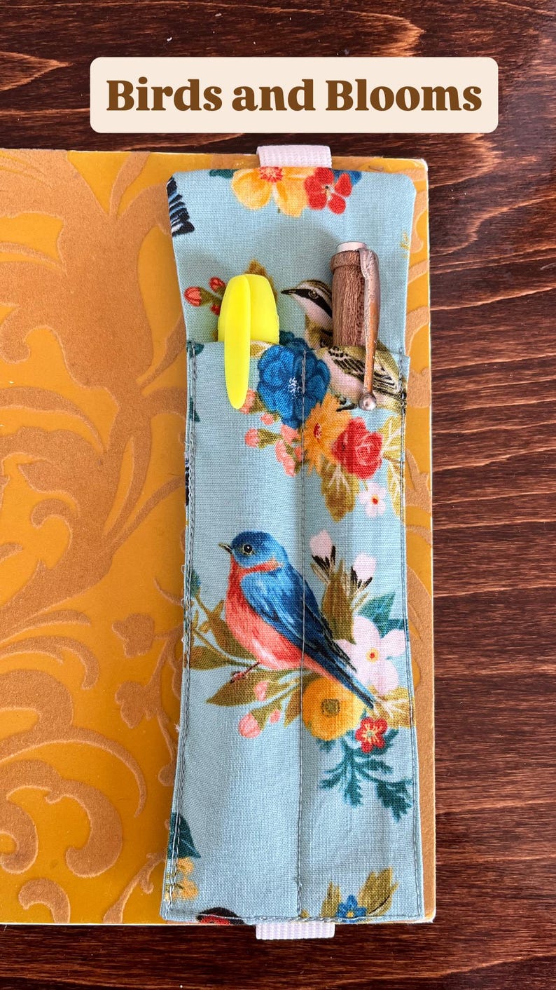 Puede incluir: Un estuche de tela azul claro con un estampado floral y de p&aacute;jaros, que contiene un rotulador amarillo y un bol&iacute;grafo de madera. El estuche est&aacute; decorado con flores y p&aacute;jaros de colores. El texto "Birds and Blooms" est&aacute; en la parte superior.