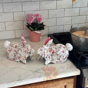 Peut inclure: Deux maniques en forme de lapin avec des motifs floraux. Les maniques sont blanches avec des fleurs rouges et roses et des feuilles vertes. Elles ont des oreilles int&eacute;rieures roses et des queues duveteuses blanches. Un petit pot de fleurs roses est derri&egrave;re.