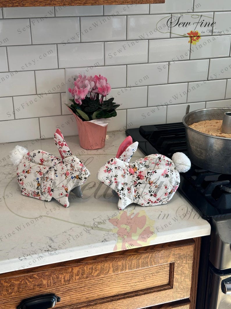 Peut inclure: Deux maniques en forme de lapin avec des motifs floraux. Les maniques sont blanches avec des fleurs rouges et roses et des feuilles vertes. Elles ont des oreilles int&eacute;rieures roses et des queues duveteuses blanches. Un petit pot de fleurs roses est derri&egrave;re.