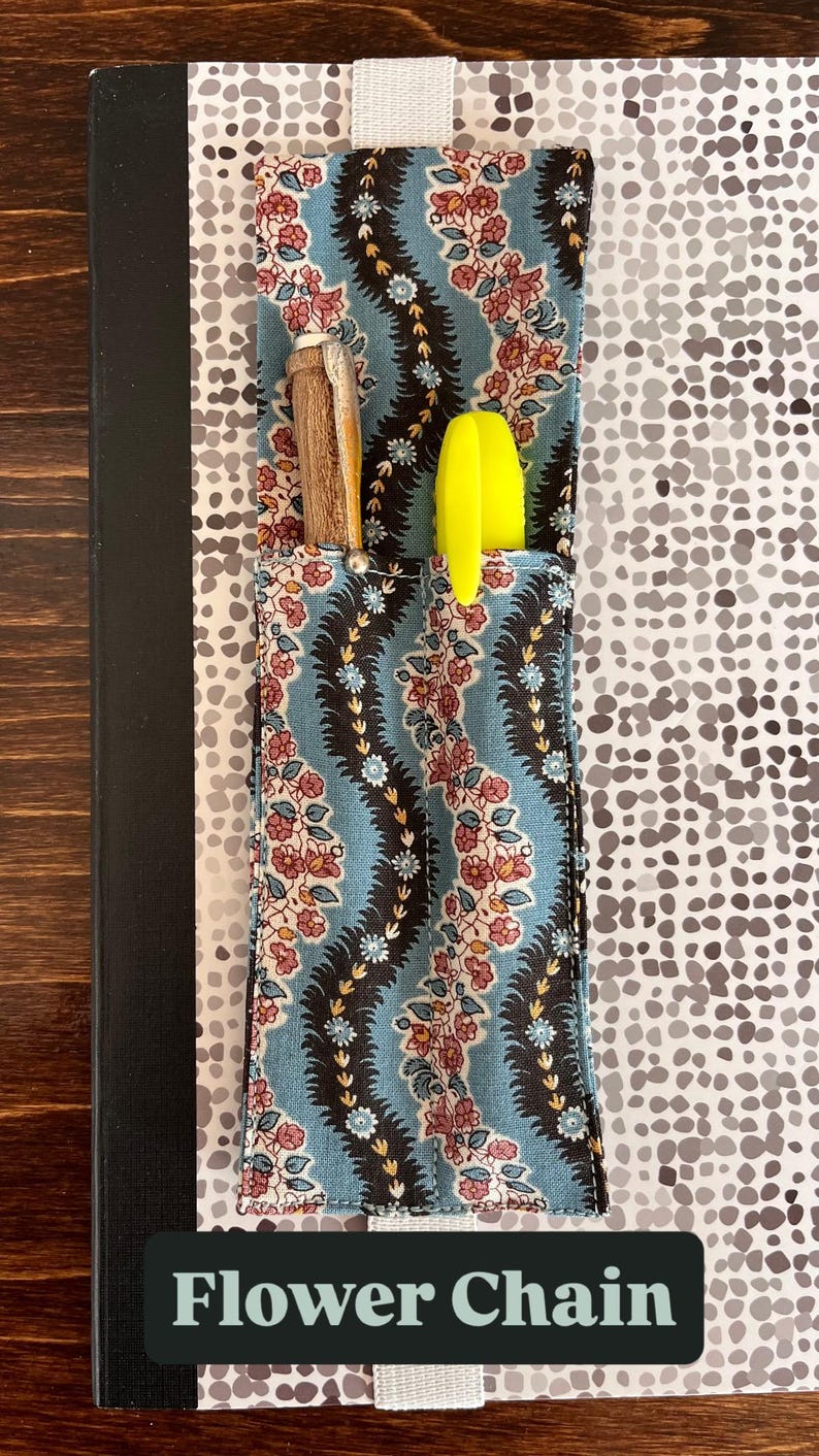 Puede incluir: Un cuaderno con un bolsillo estampado floral, que contiene un bol&iacute;grafo y un resaltador amarillo. El bolsillo tiene un fondo azul con un dise&ntilde;o floral. El texto "Flower Chain" est&aacute; en la parte inferior.