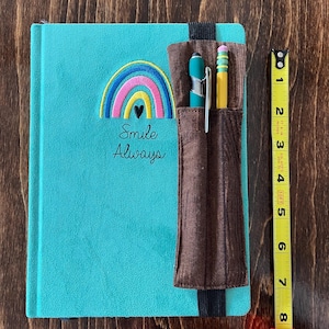 May include: A teal journal with an embroidered rainbow and the words "Smile Always." A brown pen holder is attached, holding a teal pen and a yellow pencil. A measuring tape shows the journal is approximately 8 inches long.