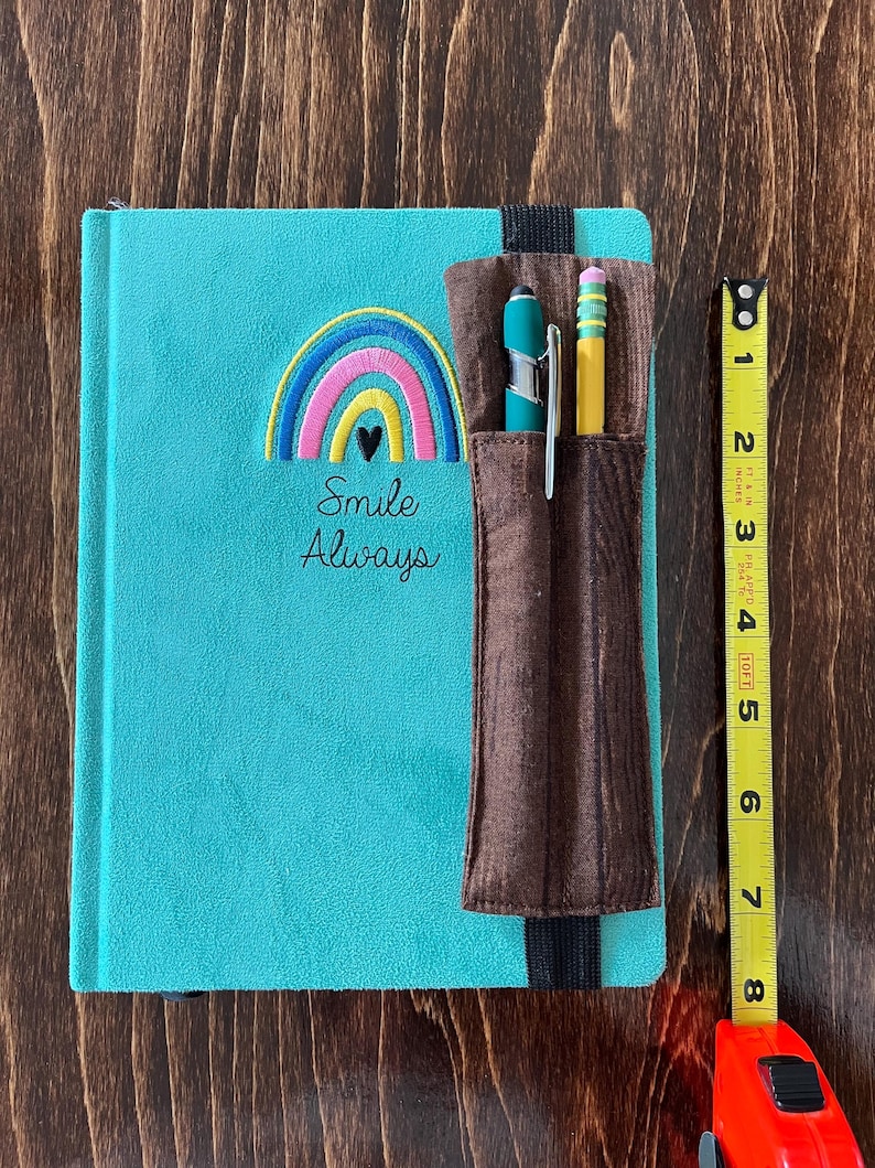 Puede incluir: Un diario turquesa con un arco&iacute;ris bordado y las palabras "Smile Always". Un portal&aacute;pices marr&oacute;n est&aacute; adjunto, con un bol&iacute;grafo turquesa y un l&aacute;piz amarillo. El diario mide aproximadamente 20 cm de largo.