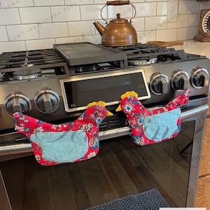 May include: Two red chicken-shaped oven mitts with blue accents and floral patterns are placed on an oven. The mitts have yellow comb details. A copper kettle and a black griddle sit on the stove top.
