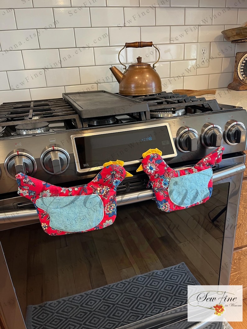 May include: Two red chicken-shaped oven mitts with blue accents and floral patterns are placed on an oven. The mitts have yellow comb details. A copper kettle and a black griddle sit on the stove top.