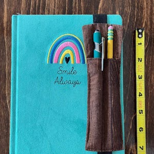 Puede incluir: Un diario turquesa con un bordado de arco&iacute;ris y coraz&oacute;n, las palabras "Smile Always", y un portal&aacute;pices marr&oacute;n. Un bol&iacute;grafo y un l&aacute;piz est&aacute;n en el soporte. El diario mide aproximadamente 20 cm de alto.