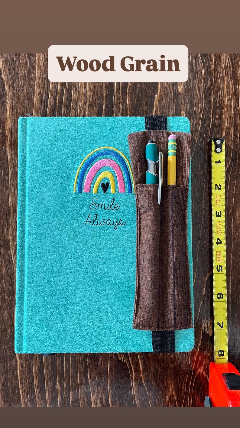 Puede incluir: Un diario turquesa con un bordado de arco&iacute;ris y coraz&oacute;n, las palabras "Smile Always", y un portal&aacute;pices marr&oacute;n. Un bol&iacute;grafo y un l&aacute;piz est&aacute;n en el soporte. El diario mide aproximadamente 20 cm de alto.