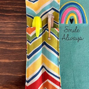 Puede incluir: Un diario turquesa con un portal&aacute;pices con estampado de zig-zag colorido. El portal&aacute;pices tiene un bolsillo con un resaltador amarillo y un bol&iacute;grafo. Un arco&iacute;ris con un coraz&oacute;n negro est&aacute; bordado en el diario, con las palabras "Smile Always".