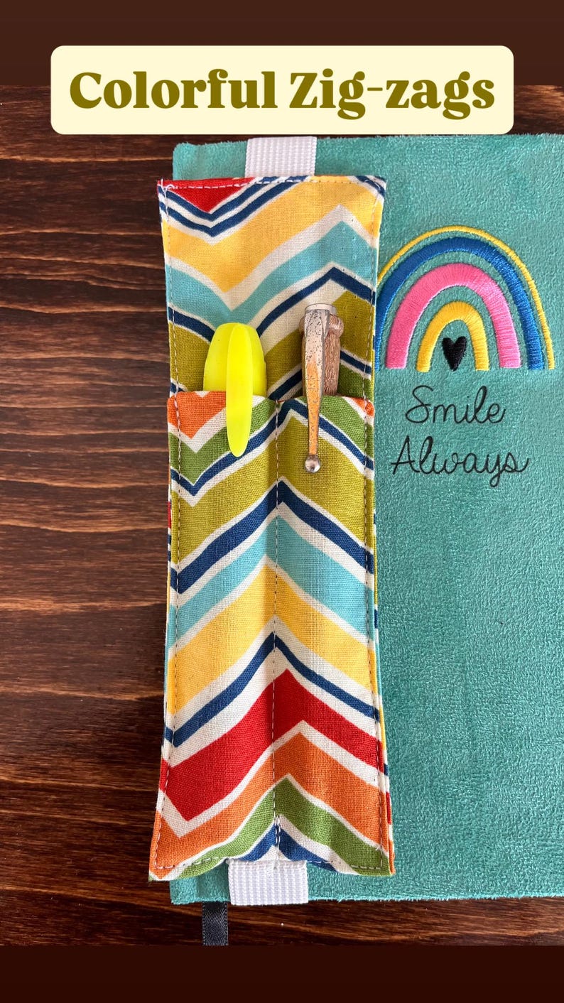 Puede incluir: Un diario turquesa con un portal&aacute;pices con estampado de zig-zag colorido. El portal&aacute;pices tiene un bolsillo con un resaltador amarillo y un bol&iacute;grafo. Un arco&iacute;ris con un coraz&oacute;n negro est&aacute; bordado en el diario, con las palabras "Smile Always".