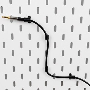 Op de afbeelding: Een zwarte audiokabel met een metalen goudkleurige jack-connector is met drie kleine zwarte kabelclips bevestigd aan een wit geperforeerd bord. De kabel loopt in een zachte bocht over het oppervlak en wordt door de clips op zijn plek gehouden.
