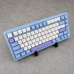May include: A mechanical keyboard with a light blue frame, featuring a mix of white, light blue, and purple keycaps. The layout includes standard QWERTY lettering, function keys, and a small logo on the right side, all supported by two black stands.
