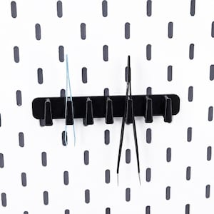 Peut inclure: Un support à outils horizontal noir doté de sept crochets régulièrement espacés, fixé sur un panneau perforé blanc à fentes ovales gris foncé. Une pince bleu clair est suspendue au deuxième crochet et une pince noire au cinquième crochet.