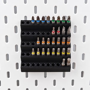 May include: A black tiered storage rack with hexagonal slots holding a variety of metal screwdriver bits. The bits feature color-coded bands in blue, green, red, orange, and yellow, organized in rows on a white pegboard background with vertical slots.
