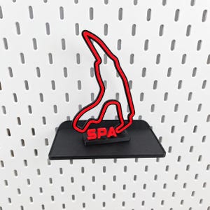 Può includere: Una mensola nera montata a parete, caratterizzata da un contorno rosso del circuito di Spa-Francorchamps. La parola SPA è stampata in lettere rosse a blocchi alla base del tracciato, centrato sulla superficie nera piatta.