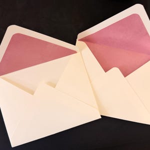 May include: Two open cream-coloured envelopes with pointed flaps. The interior of each envelope is lined with a solid, matte mauve-pink paper insert. The envelopes are positioned side-by-side, displaying the triangular flap and the inner lining.