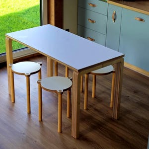 May include: A modern dining table with a white tabletop and light wood frame, paired with three matching round stools. The table is rectangular, and the stools have white seats and light wood legs. The room has a light blue cabinet and a large window.