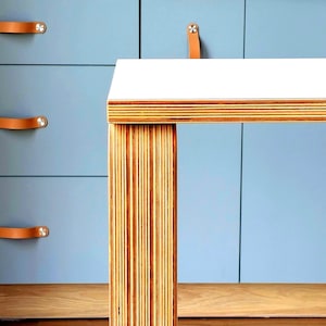 May include: A table with a white rectangular top and a thick leg constructed from layered plywood, showcasing the visible wood grain edges. The table features a clean, flat surface and a sturdy, vertically oriented support structure made of stacked wood.