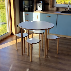Puede incluir: Una mesa redonda blanca con cuatro taburetes a juego. La mesa y los taburetes tienen patas de madera clara y tapas blancas. La mesa tiene aproximadamente 91 cm de diámetro. Los taburetes son pequeños y redondos, adecuados para un área de juegos infantil o un comedor pequeño.