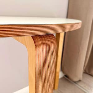 Peut inclure: Une table ronde dotée d'un plateau blanc lisse et d'un pied incurvé en contreplaqué stratifié. Le bord du plateau et le pied révèlent le motif distinctif et empilé des couches de placage en bois clair.