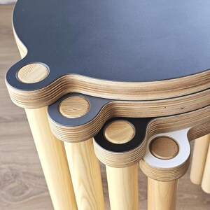 May include: A set of three stacked round stools featuring layered plywood edges with visible wood grain. Each stool has a flat top surface in black or white, with a protruding circular tab detail that aligns vertically with light wood legs.