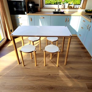Op de afbeelding: Een witte rechthoekige tafel met vier bijpassende krukken met ronde bovenkant. De tafel en krukken hebben lichtgekleurde houten poten en witte oppervlakken. De meubels staan in een keuken met lichtblauwe kasten en houten vloeren.