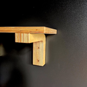 May include: A wooden shelf with a right-angle bracket, mounted on a dark wall. The shelf and bracket are made of layered plywood, with visible wood grain. Two screws secure the bracket to the wall, showcasing a minimalist design.