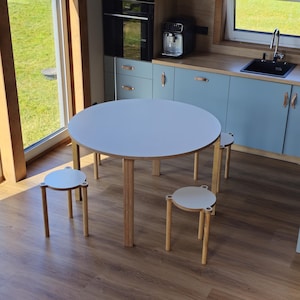 Puede incluir: Una mesa de comedor redonda blanca con patas de madera clara y tres taburetes redondos a juego con patas de madera clara. La mesa y los taburetes tienen una superficie blanca lisa con bordes de madera clara, sobre un suelo de madera.