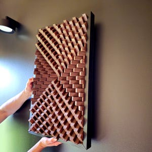 Pode incluir: Uma peça de arte de parede retangular de madeira com um design geométrico. A obra de arte apresenta uma série de pequenos blocos retangulares dispostos em um padrão repetitivo, criando uma superfície texturizada. A moldura é cinza escuro e os blocos são marrons quentes.