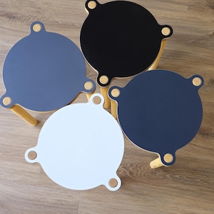 May include: Four circular stools with light-colored wooden legs and tops in black, white, and two shades of blue. Each stool features three protruding tabs with visible wooden circular accents at the connection points to the legs.