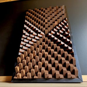 Może przedstawiać: Geometryczna drewniana grafika ścienna z ciemną, bejcowaną ramą. Grafika przedstawia wzór małych, prostokątnych bloków w różnych odcieniach brązu, tworząc teksturowany, trójwymiarowy efekt. Projekt jest kątowy, dodając wizualnego zainteresowania.