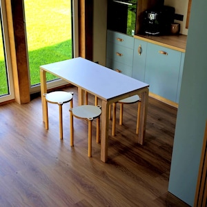 May include: A modern dining set featuring a rectangular table with a white top and light wood frame. Three matching round stools with white seats and light wood legs are positioned around the table. The set is in a room with a wooden floor and a view of green grass.