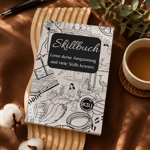 Könnte beinhalten: Ein weißes Skillbuch-Notizbuch mit schwarzen Strichzeichnungen und Text, das auf einem Holztablett liegt. Das Cover des Notizbuchs trägt den Titel "Skillbuch" und deutschen Text. Ein Stift, eine Tasse Tee und Baumwollzweige sind ebenfalls im Bild.