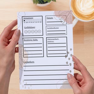 Könnte beinhalten: Ein weißes Papier mit deutschem Text, das von Händen gehalten wird und Abschnitte zur Verfolgung von Stimmung, Fähigkeiten und Gedanken enthält. Das Papier enthält eine Stresspegelanzeige und Platz für ein Datum. Eine Tasse Kaffee und eine Pflanze befinden sich im Hintergrund.