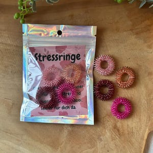 Könnte beinhalten: Eine Packung Stressringe in einer holografischen Tüte, mit dem Wort "Stressringe" sichtbar. Mehrere Ringe sind um die Tüte herum verstreut, in Rosa- und Kupfertönen. Die Ringe bestehen aus kleinen, stacheligen Metallspiralen.