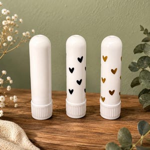 Könnte beinhalten: Drei weiße, zylindrische Inhalator-Stifte, die aufrecht stehen. Der erste Stift ist schlicht weiß, der zweite ist mit kleinen schwarzen Herzen bedruckt und der dritte mit kleinen metallisch goldenen Herzen. Jeder Stift hat eine geriffelte Basis.