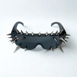 Puede incluir: Gafas de sol negras con púas plateadas en los bordes superior e inferior. Las gafas tienen una montura curvada y lentes oscuras. Las púas están espaciadas uniformemente y añaden una estética audaz y vanguardista.