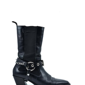 May include: Black leather cowboy boots with a silver chain and buckle detail. The boots have a pointed toe and a block heel.