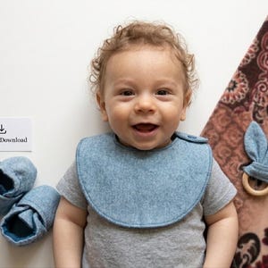 May include: A smiling baby wearing a blue bib and gray shirt, with matching blue booties and a bunny ear teether. A sign reads "PDF Bib Download". The items are on a white surface with a patterned blanket.