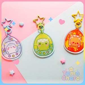 May include: Three colorful acrylic keychains with cartoon animal designs. The keychains feature a penguin, a frog, and a bear, each inside a stylized egg-shaped frame. The keychains have star-shaped clasps and are set against a pink and blue background.