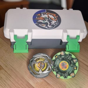 Può includere: Una custodia rettangolare bianca e grigia con due chiusure in plastica verde brillante e un grafico circolare di una tigre sul coperchio. Due trottole sono posizionate davanti alla custodia: una con una cornice metallica argentata e centro giallo, e una con un design a ingranaggio verde scuro e un emblema circolare centrale.