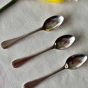 May include: Three silver spoons with beaded edges are arranged on a white surface. A yellow daffodil and a white lace doily are in the background. The spoons have a reflective surface.
