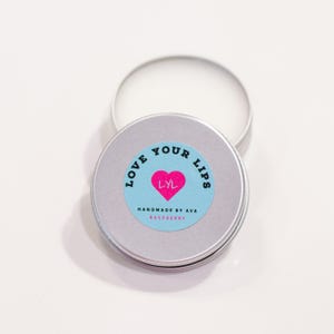 Könnte beinhalten: Eine silberne Dose mit einem blauen und weißen Aufkleber, auf dem "Love Your Lips" mit einem rosa Herzen und den Buchstaben "LYL" darin steht. Der Aufkleber sagt auch "Handmade by Ava" und "Raspberry".