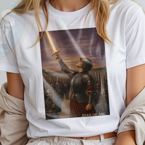 May include: White t-shirt featuring a print of Joan of Arc holding a sword aloft, with the text "JOAN OF ARC" below the image. The shirt has short sleeves and a crew neck.