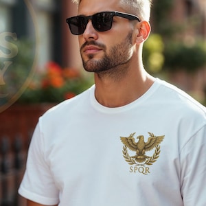 May include: White t-shirt featuring a gold emblem of an eagle with a laurel wreath and the letters SPQR. The shirt is worn by a person wearing sunglasses. The image also includes the text "TEE VIBES ITALY".